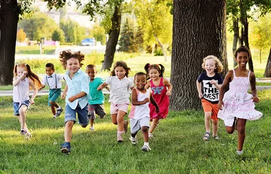 Neun Kinder im Kindergarten und Grundschulalter rennen mit fliegenden Haaren um die Wette – sie laufen auf die Kamera zu. Sie sind sommerlich in bunten T-Shirts, Shirts und Kleidern gekleidet.