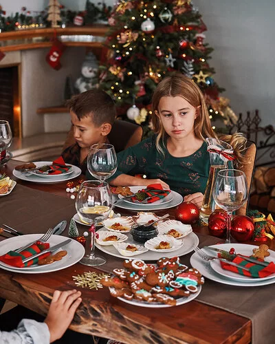 Eine Frau sitzt mit vier Kindern an einem weihnachtlich gedeckten Tisch, auf dem Plätzchen stehen. Zwei der Kinder sehen betrübt aus.