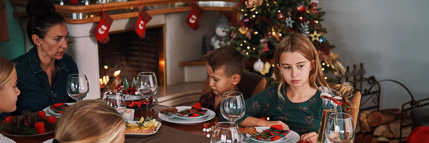 Eine Frau sitzt mit vier Kindern an einem weihnachtlich gedeckten Tisch, auf dem Plätzchen stehen. Zwei der Kinder sehen betrübt aus.