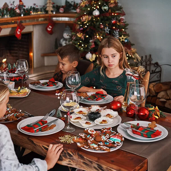 Eine Frau sitzt mit vier Kindern an einem weihnachtlich gedeckten Tisch, auf dem Plätzchen stehen. Zwei der Kinder sehen betrübt aus.