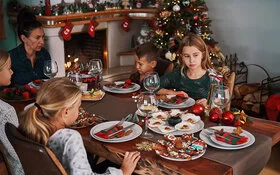 Eine Frau sitzt mit vier Kindern an einem weihnachtlich gedeckten Tisch, auf dem Plätzchen stehen. Zwei der Kinder sehen betrübt aus.