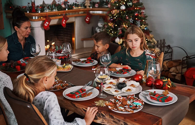 Eine Frau sitzt mit vier Kindern an einem weihnachtlich gedeckten Tisch, auf dem Plätzchen stehen. Zwei der Kinder sehen betrübt aus.