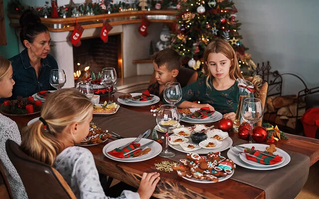 Eine Frau sitzt mit vier Kindern an einem weihnachtlich gedeckten Tisch, auf dem Plätzchen stehen. Zwei der Kinder sehen betrübt aus.