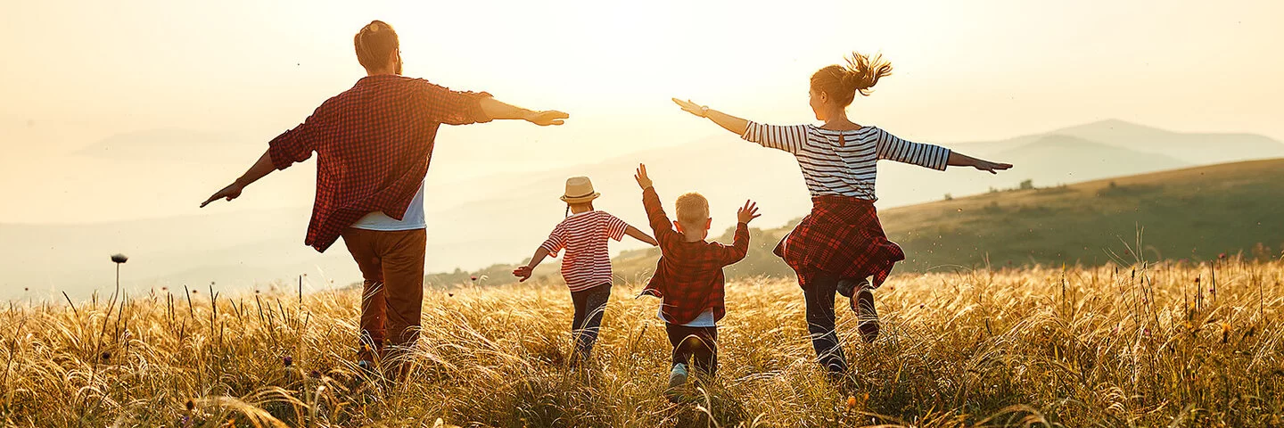 Mutter, Vater, Sohn und Tochter laufen bei Sonnenuntergang mit ausgestreckten Armen über eine Wiese mit hohen Gräsern in Richtung Sonnenuntergang.