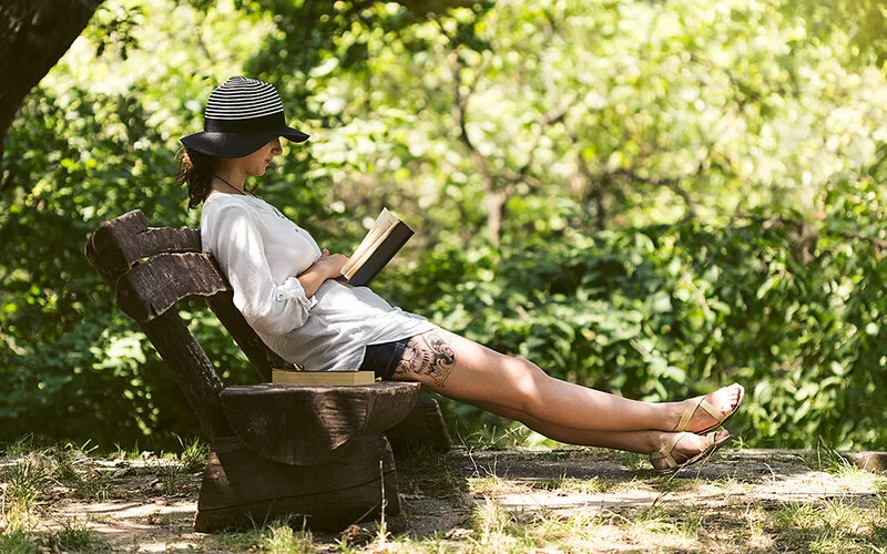 Eine Frau mit Sonnenhut sitzt auf einer Bank unter dichten Bäumen und liest ein Buch.