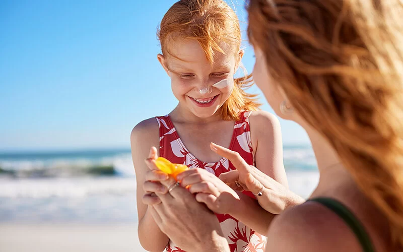 Eine Mutter hilft ihrer Tochter, sich am Strand mit Sonnencreme einzucremen.