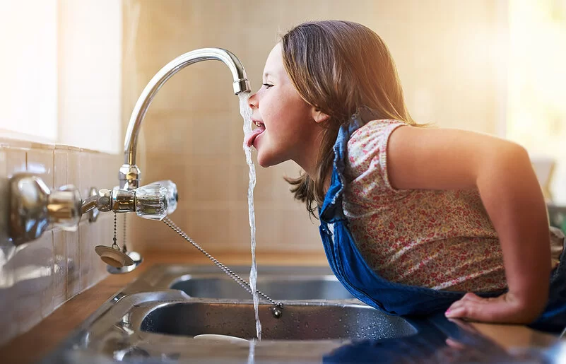 Ein Mädchen beugt sich über die Spüle und streckt die Zunge nach dem Wasser aus dem Wasserhahn aus.