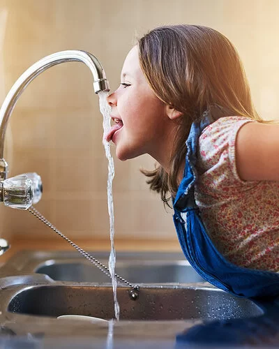 Ein Mädchen beugt sich über die Spüle und streckt die Zunge nach dem Wasser aus dem Wasserhahn aus.