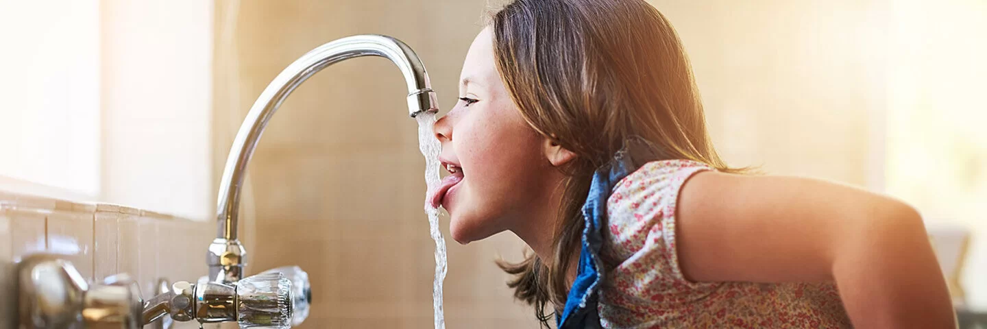 Ein Mädchen beugt sich über die Spüle und streckt die Zunge nach dem Wasser aus dem Wasserhahn aus.