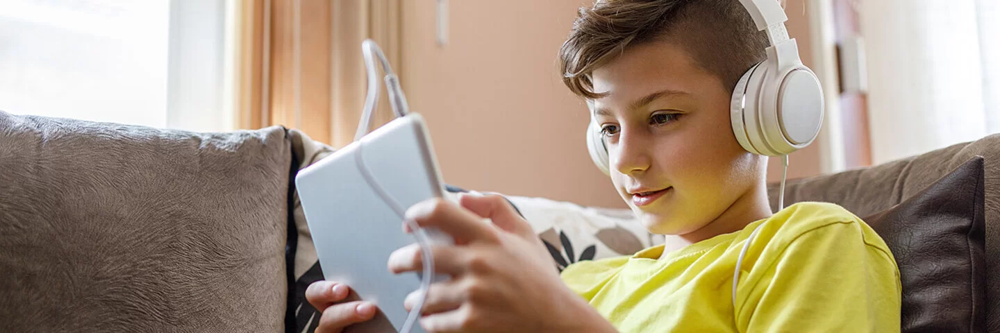 Ein Junge sitzt mit einem Tablet und Kopfhörern auf einem Sofa und schaut auf den Bildschirm.