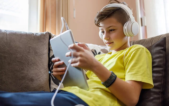 Ein Junge sitzt mit einem Tablet und Kopfhörern auf einem Sofa und schaut auf den Bildschirm.