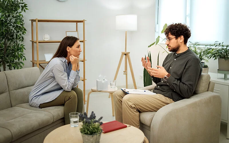 Eine junge Frau sitzt auf einem Sofa im Sprechzimmer eines Psychotherapeuten. Der Therapeut sitzt ihr mit einem Klemmbrett auf dem Schoß in einem Sessel gegenüber.