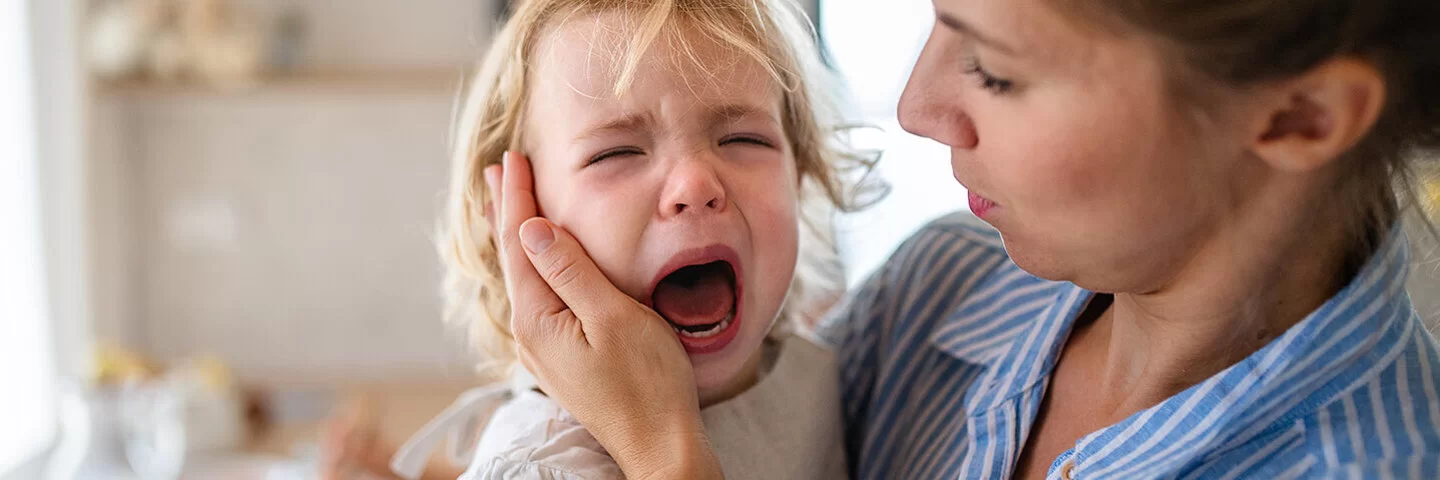 Eine Mutter hält in der Küche ihre weinende Kleinkindtochter auf dem Arm und streicht ihr zärtlich über die Wange.