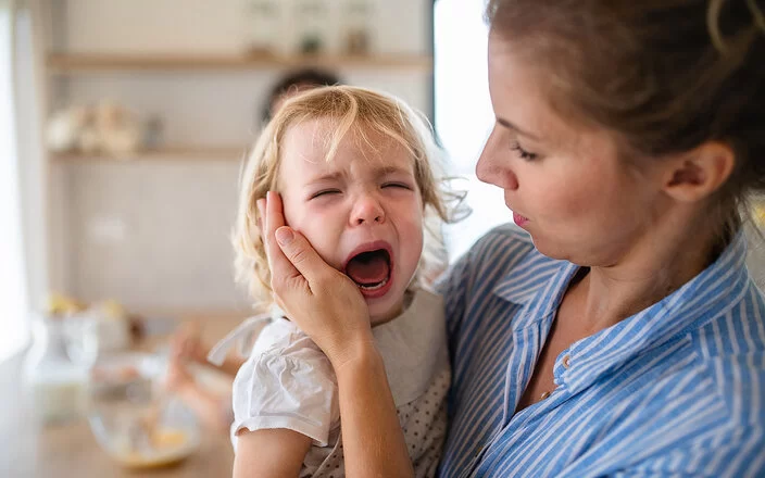 Eine Mutter hält in der Küche ihre weinende Kleinkindtochter auf dem Arm und streicht ihr zärtlich über die Wange.