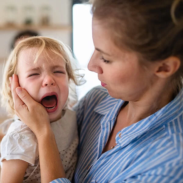 Eine Mutter hält in der Küche ihre weinende Kleinkindtochter auf dem Arm und streicht ihr zärtlich über die Wange.