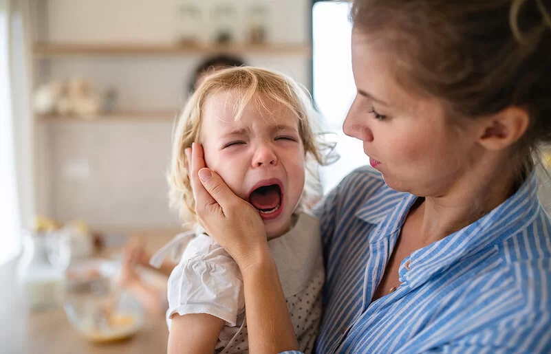 Eine Mutter hält in der Küche ihre weinende Kleinkindtochter auf dem Arm und streicht ihr zärtlich über die Wange.