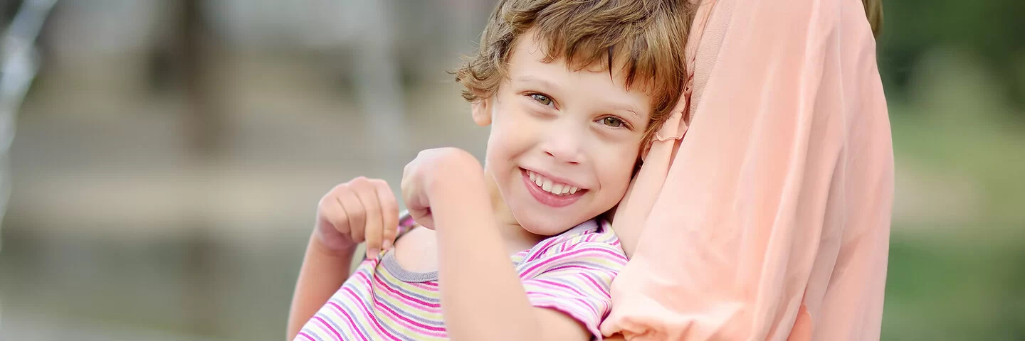 Ein durch eine Zerebralparese beeinträchtigtes Mädchen sitzt auf dem Schoß seiner Mutter im Freien. Das Mädchen trägt einen kurzärmeligen Body und lächelt in die Kamera, während die Mutter, von der nur der Oberkörper abgebildet ist, das Kind umarmt.