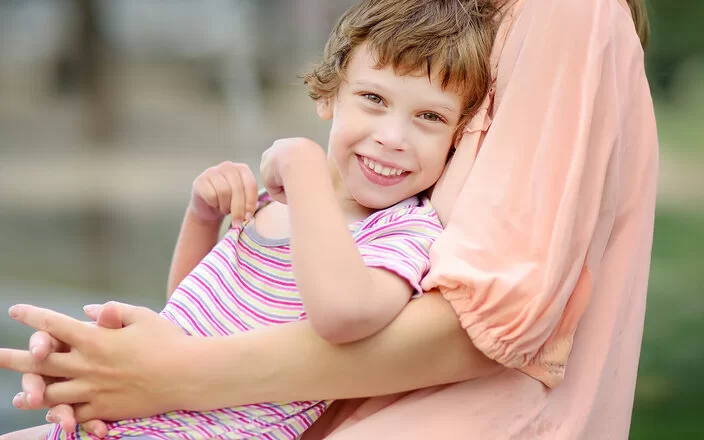 Ein durch eine Zerebralparese beeinträchtigtes Mädchen sitzt auf dem Schoß seiner Mutter im Freien. Das Mädchen trägt einen kurzärmeligen Body und lächelt in die Kamera, während die Mutter, von der nur der Oberkörper abgebildet ist, das Kind umarmt.