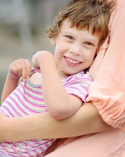 Ein durch eine Zerebralparese beeinträchtigtes Mädchen sitzt auf dem Schoß seiner Mutter im Freien. Das Mädchen trägt einen kurzärmeligen Body und lächelt in die Kamera, während die Mutter, von der nur der Oberkörper abgebildet ist, das Kind umarmt.