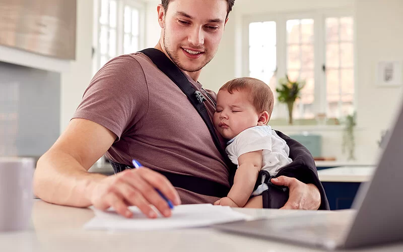 Ein junger Mann sitzt zu Hause an seinem Schreibtisch und füllt ein Formular aus. Vor seiner Brust hält er sein Baby in einer Trage.
