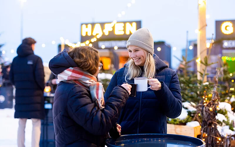 Zwei Frauen stehen draußen auf einem winterlichen Weihnachtsmarkt und halten Tassen in den Händen. Eine junge Frau mit blonden Haaren und beiger Mütze lächelt ihre Gesprächspartnerin an, die einen karierten Schal trägt, beide tragen warme schwarze Jacken. Im Hintergrund sind Lichterketten, Tannenbäume und ein leuchtendes Schild mit der Aufschrift „Hafen“ zu sehen, die eine gemütliche, festliche Atmosphäre schaffen.