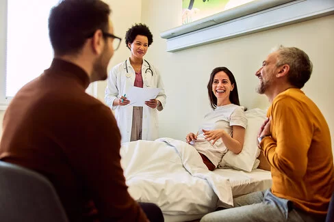 Eine jüngere Frau sitzt aufgerichtet auf einem Krankenhausbett, rechts von ihr steht eine Ärztin mit Unterlagen in den Händen, links vorm Bett sitzen zwei Männer, alle lachen.