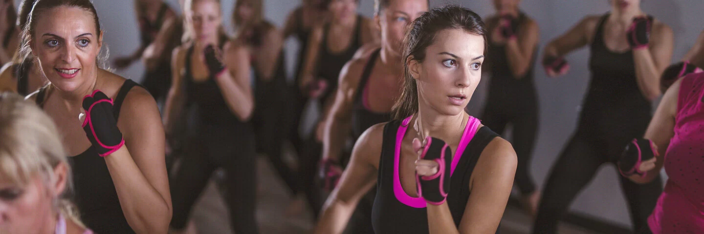 Im Fokus sind zwei Frauen in Box-Pose zu sehen, 17 weitere Frauen in der Unschärfe. Die gesamte Piloxing-Gruppe trägt fingerlose, schwarz-pinke Handschuhe und überwiegend schwarz-pinke Sportkleidung.