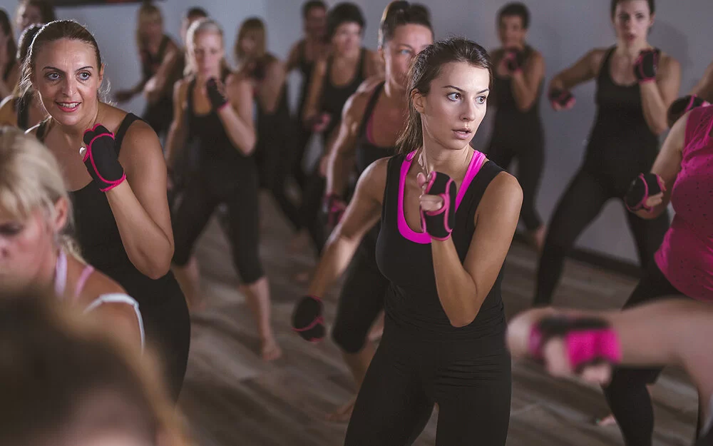 Im Fokus sind zwei Frauen in Box-Pose zu sehen, 17 weitere Frauen in der Unschärfe. Die gesamte Piloxing-Gruppe trägt fingerlose, schwarz-pinke Handschuhe und überwiegend schwarz-pinke Sportkleidung.