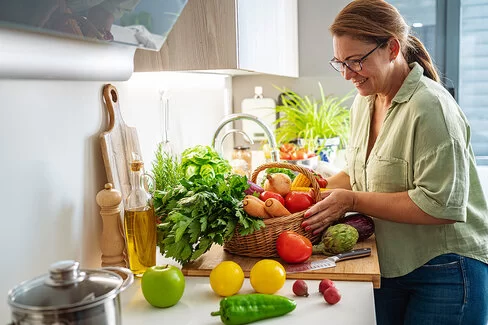 Eine Frau steht in der Küche und packt ihren Einkaufskorb aus. Vor ihr liegt zahlreiches frisches Obst und Gemüse auf der Arbeitsfläche wie ein grüner Apfel, zwei Zitronen, eine grüne Paprika, mehrere Radieschen, eine Tomate und eine Artischocke. In ihrem Korb liegen noch Petersilie, Möhren und Zwiebeln. 