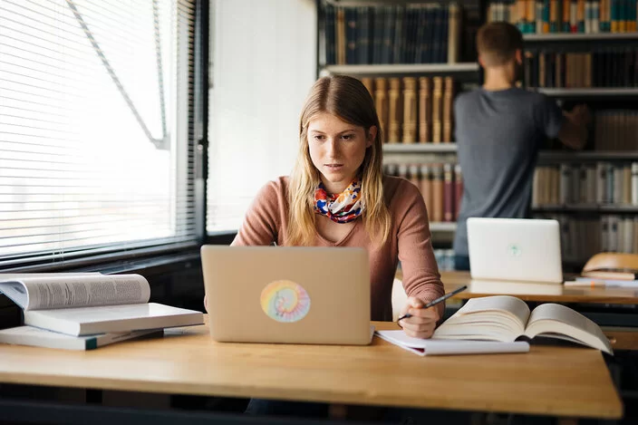 Eine junge Frau sitzt an einem Tisch und schaut auf einen Laptop. Auf dem Tisch liegen ein paar aufgeschlagene Bücher.