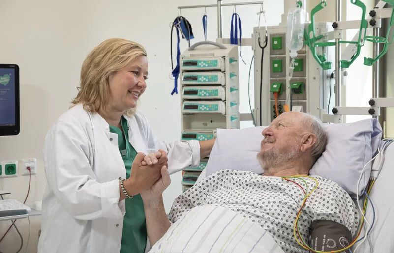 Auf dem Bild ist ein älterer Mann in einem Krankenhausbett zu sehen. Neben ihm steht eine Ärztin, die ihm die Hand hält. 