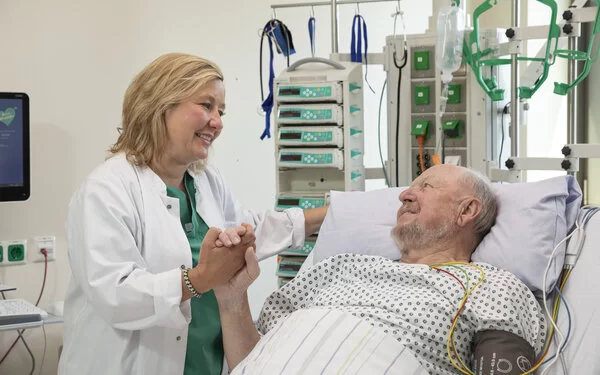 Auf dem Bild ist ein älterer Mann in einem Krankenhausbett zu sehen. Neben ihm steht eine Ärztin, die ihm die Hand hält. 