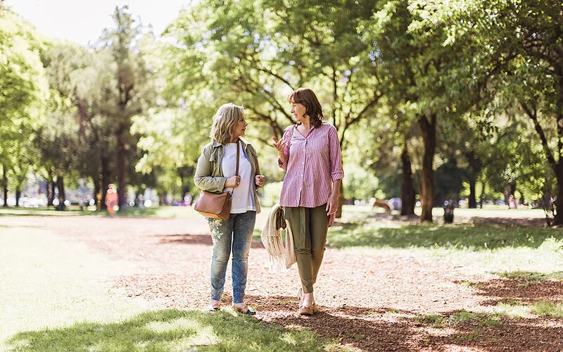 Zwei Frauen um die 50 gehen in einem sommerlichen Park spazieren und unterhalten sich.