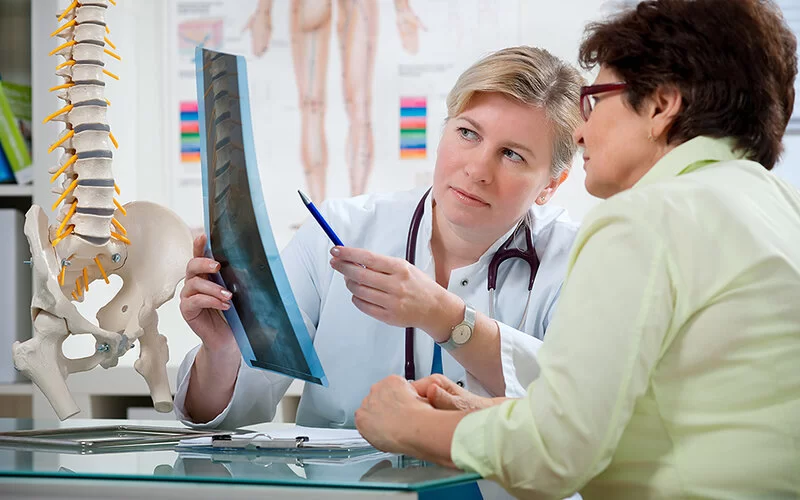 Eine Orthopädin erläutert in ihrem Sprechzimmer einer älteren Patientin das Röntgenbild von deren Wirbelsäule.