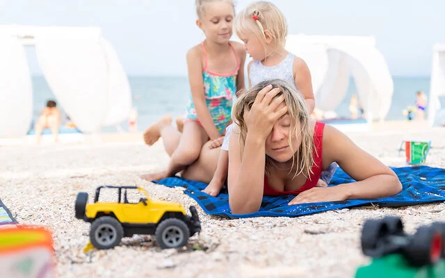 Eine Frau liegt mit dem Bauch auf einem Handtuch am Strand und fasst sich mit einer Hand an die Stirn. Zwei kleine Kinder sitzen auf ihrem Rücken.