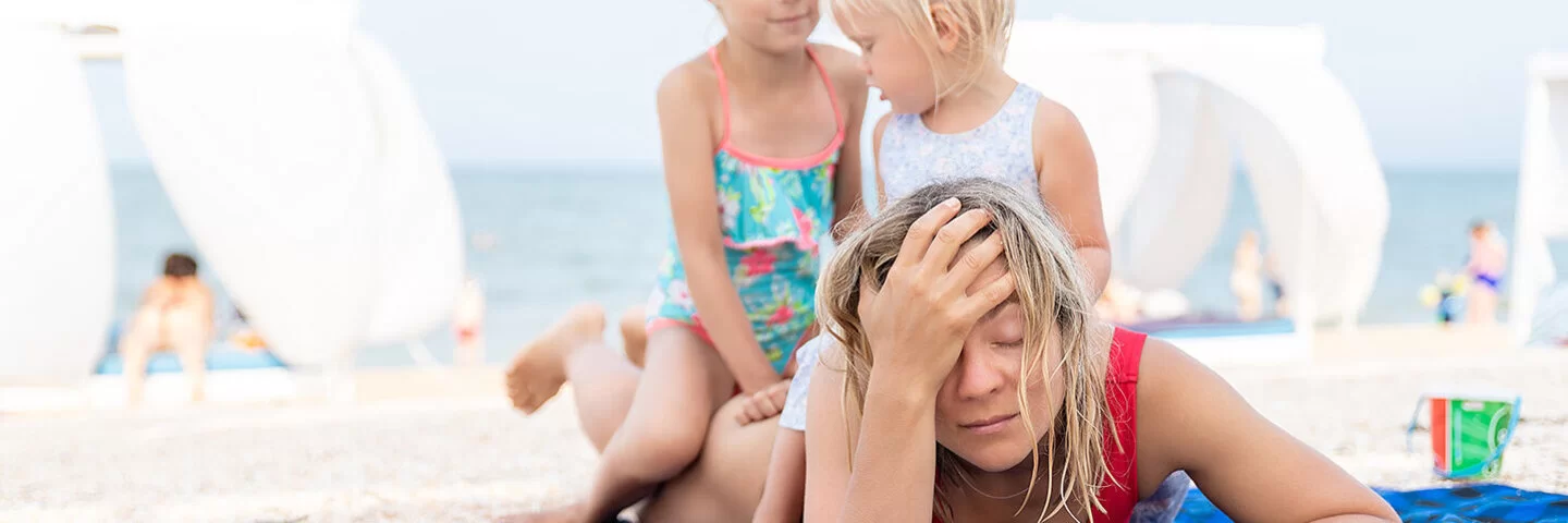 Eine Frau liegt mit dem Bauch auf einem Handtuch am Strand und fasst sich mit einer Hand an die Stirn. Zwei kleine Kinder sitzen auf ihrem Rücken.