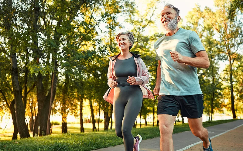 Eine ältere Frau und ein älterer Mann joggen in einem sommerlichen Park. Durch die Bäume im Hintergrund schimmern die Strahlen der aufgehenden Sonne.