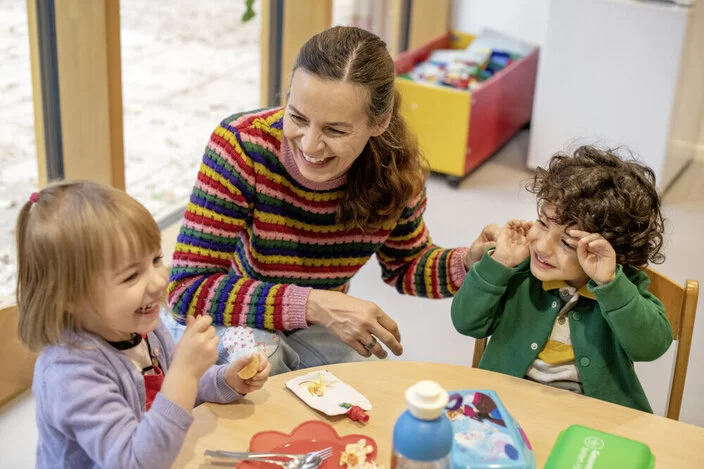 Zwei Kinder sitzen im Kindergarten an einem niedrigen Tisch. Das Kind links im Bild trägt einen violetten Pullover und isst lächelnd. Das Kind rechts im Bild trägt eine dunkelgrüne Jacke und hat seine Hände neben seinem Gesicht. Zwischen ihnen hockt die Erzieherin lächelnd in einem bunt gestreiften Pullover.