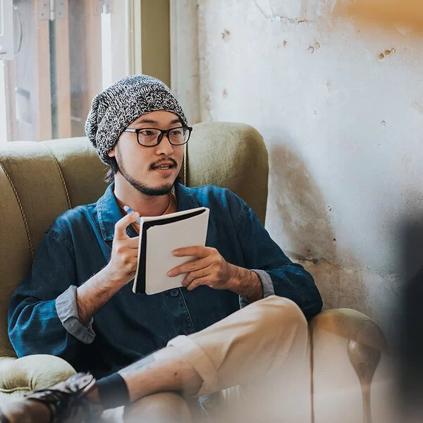 Ein Mann sitzt mit überschlagenen Beinen auf einem Sessel. Er hält einen Stift und ein Notizbuch in der Hand.