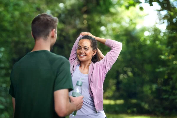 Eine junge Frau in den Zwanzigern steht in einem Park und mach Dehnübungen mit den Armen. Ihr gegenüber steht ein nicht erkennbarer Mann mit einer Trinkflasche in der rechten Hand.