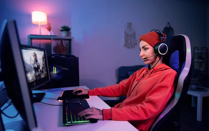 Eine junge Frau sitzt mit Kopfhörern vor mehreren nebeneinander platzierten Monitoren und spielt ein Computerspiel.