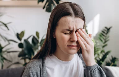 Eine junge Frau sitzt mit schmerzerfülltem Gesicht in einem Wohnzimmer. Ihre Augen sind geschlossen. Mit der linken Hand fasst sie sich an das linke Auge.