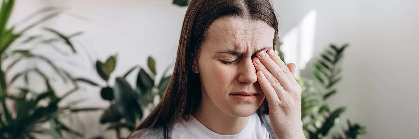 Eine junge Frau sitzt mit schmerzerfülltem Gesicht in einem Wohnzimmer. Ihre Augen sind geschlossen. Mit der linken Hand fasst sie sich an das linke Auge.