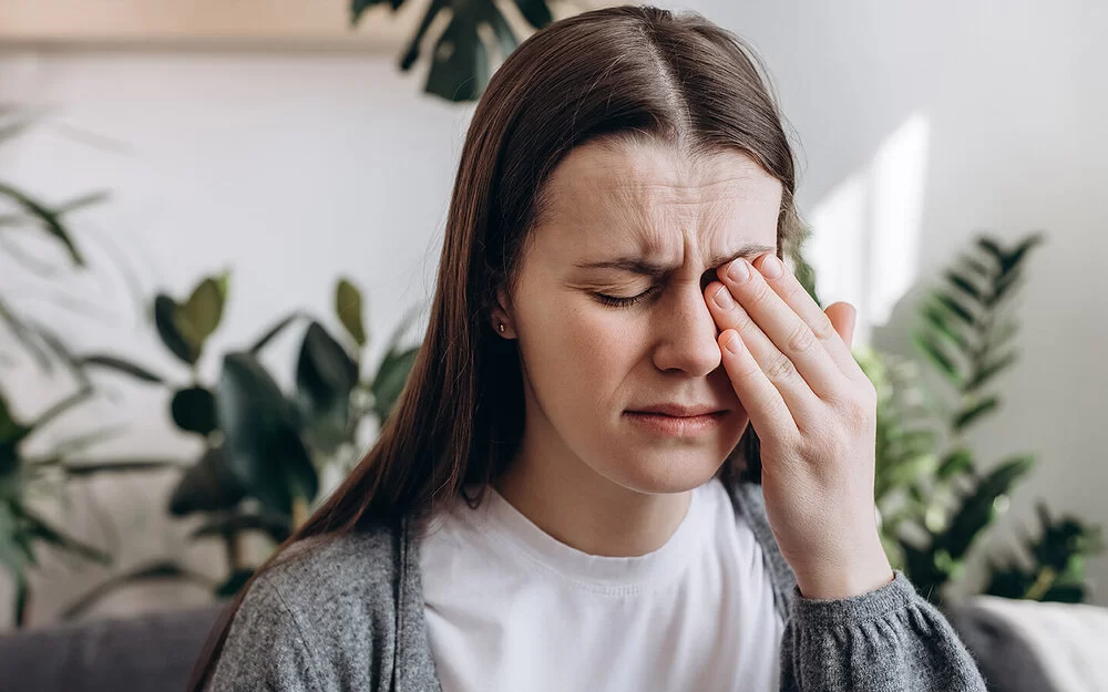 Eine junge Frau sitzt mit schmerzerfülltem Gesicht in einem Wohnzimmer. Ihre Augen sind geschlossen. Mit der linken Hand fasst sie sich an das linke Auge.