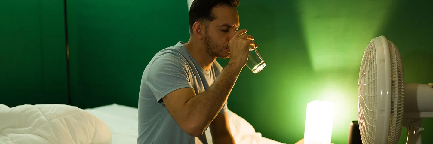 Mann sitzt auf der Bettkante und trinkt ein Glas Wasser, weil er bei Hitze nicht schlafen kann.