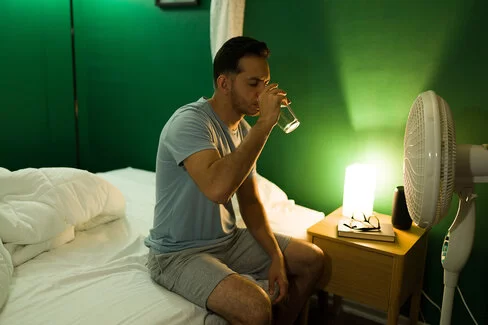 Mann sitzt auf der Bettkante und trinkt ein Glas Wasser, weil er bei Hitze nicht schlafen kann.