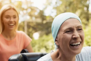 Eine lachende Frau schiebt eine Lungenkrebs-Patientin im Rollstuhl, die glücklich die Arme nach oben streckt.