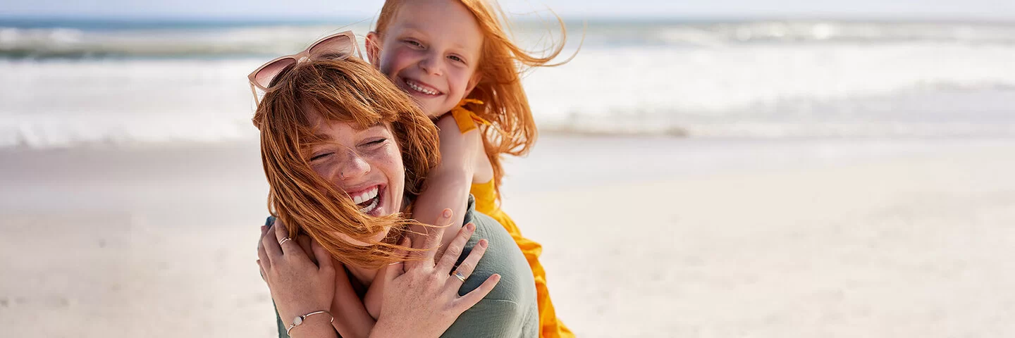 Eine junge Frau trägt ein Mädchen Huckepack an einem Strand, beide lachen fröhlich, der Wind weht ihnen das Haar ins Gesicht.