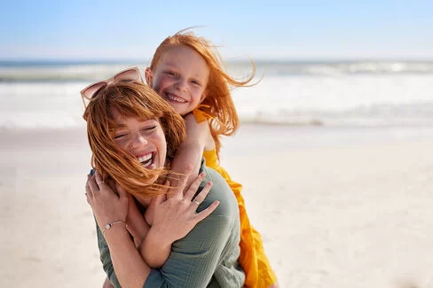 Eine junge Frau trägt ein Mädchen Huckepack an einem Strand, beide lachen fröhlich, der Wind weht ihnen das Haar ins Gesicht.