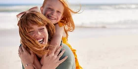 Eine junge Frau trägt ein Mädchen Huckepack an einem Strand, beide lachen fröhlich, der Wind weht ihnen das Haar ins Gesicht.